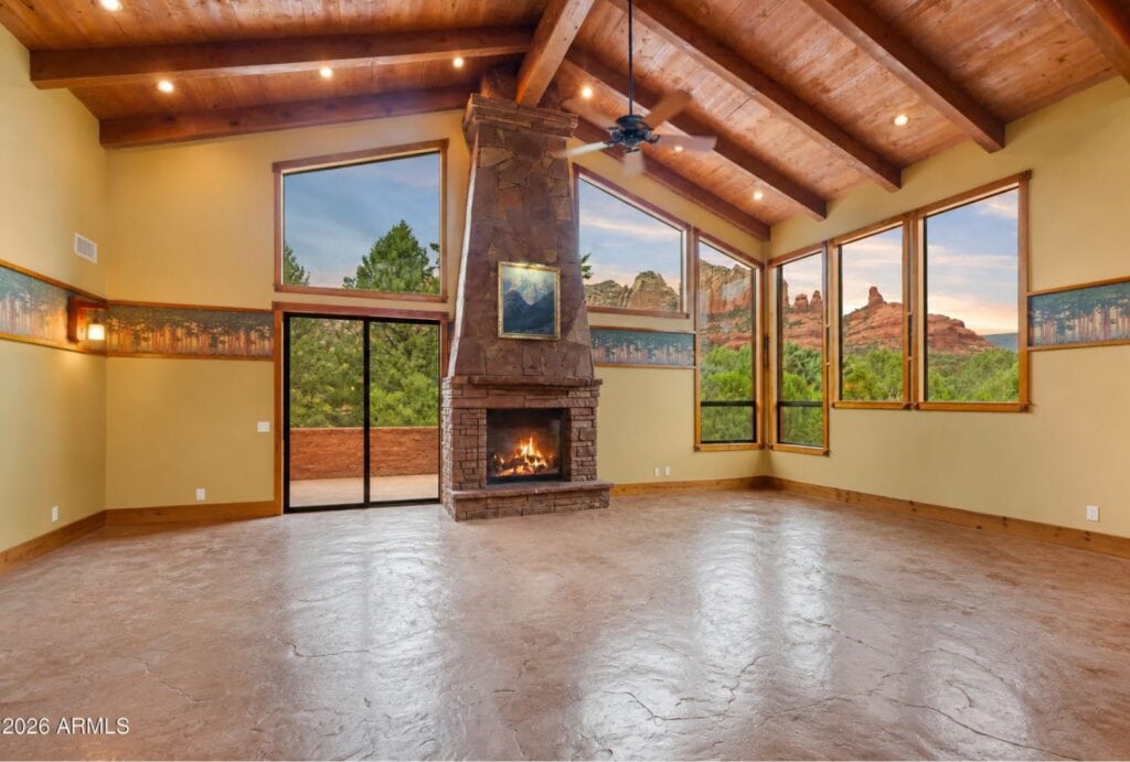A spacious living room featuring a tall stone fireplace, wood-beamed ceilings, and large windows overlooking Sedona red rocks, serving as inspiration for biophilic design by Sedona Interior Designer Dana Cavanaugh.
